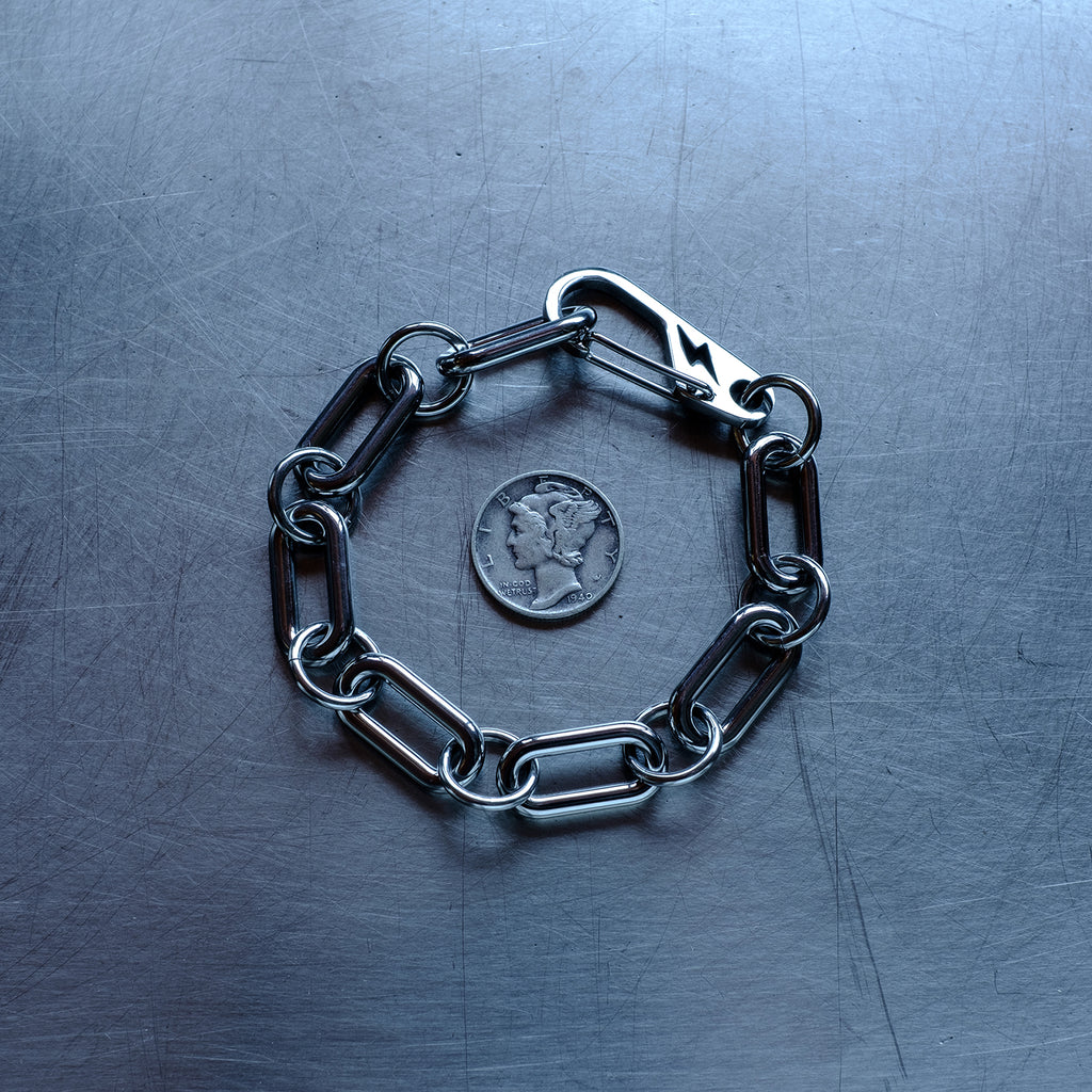 Statement, chunky Oval chain link connected by large round jump rings bracelet featuring a carabiner clasp with a lightning bolt cutout made with stainless steel on a stainless steel surface with a Liberty Dime for scale