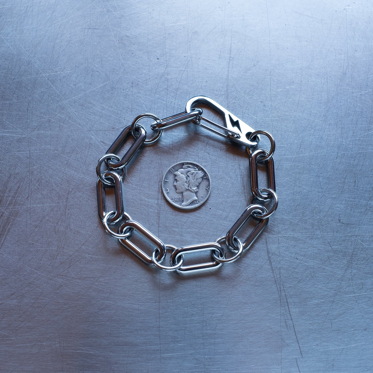 Statement, chunky Oval chain link connected by large round jump rings bracelet featuring a carabiner clasp with a lightning bolt cutout made with stainless steel on a stainless steel surface with a Liberty Dime for scale