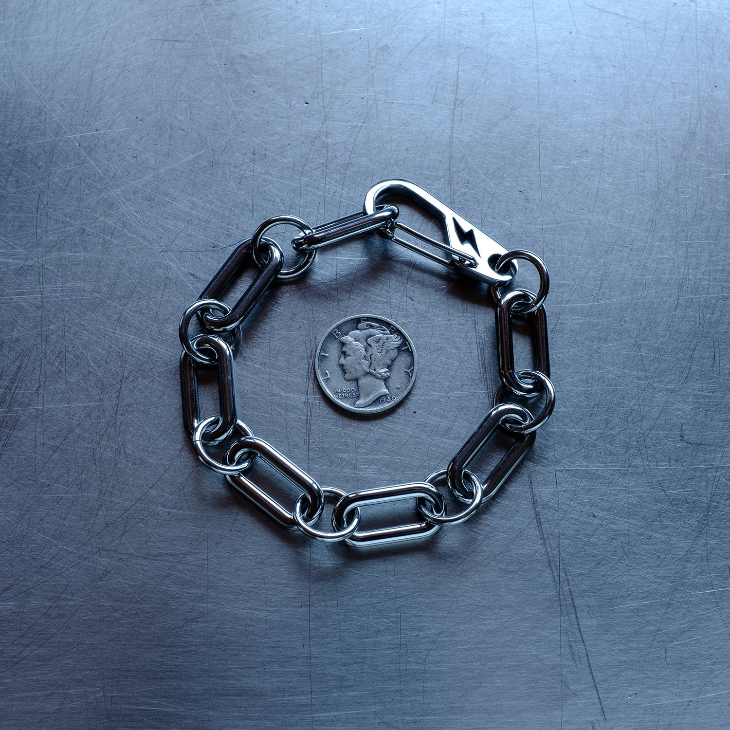 Statement, chunky Oval chain link connected by large round jump rings bracelet featuring a carabiner clasp with a lightning bolt cutout made with stainless steel on a stainless steel surface with a Liberty Dime for scale